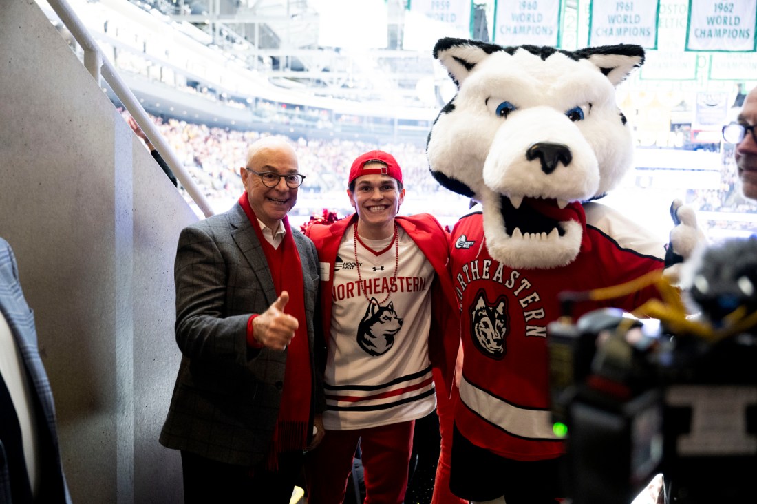 A Northeastern mascot posing with a fan in a jersey and an older man giving a thumbs-up in front of a brightly lit arena.