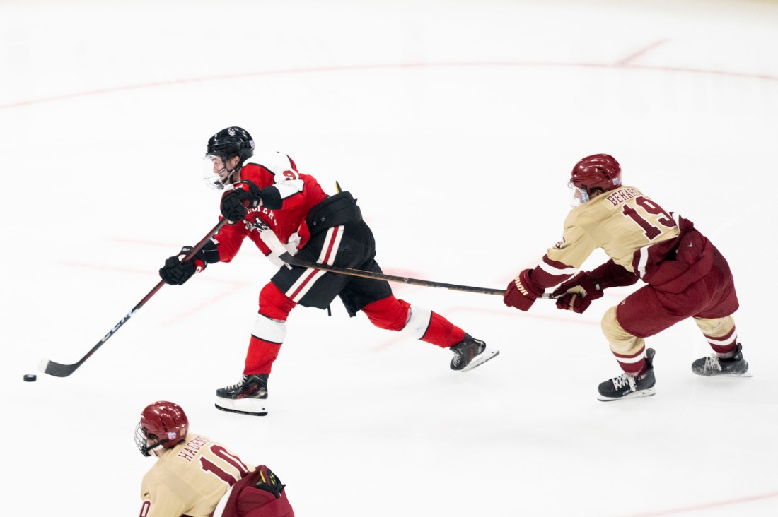 A hockey player in a red and black jersey skating with the puck while an opposing player in cream and maroon chases closely behind.