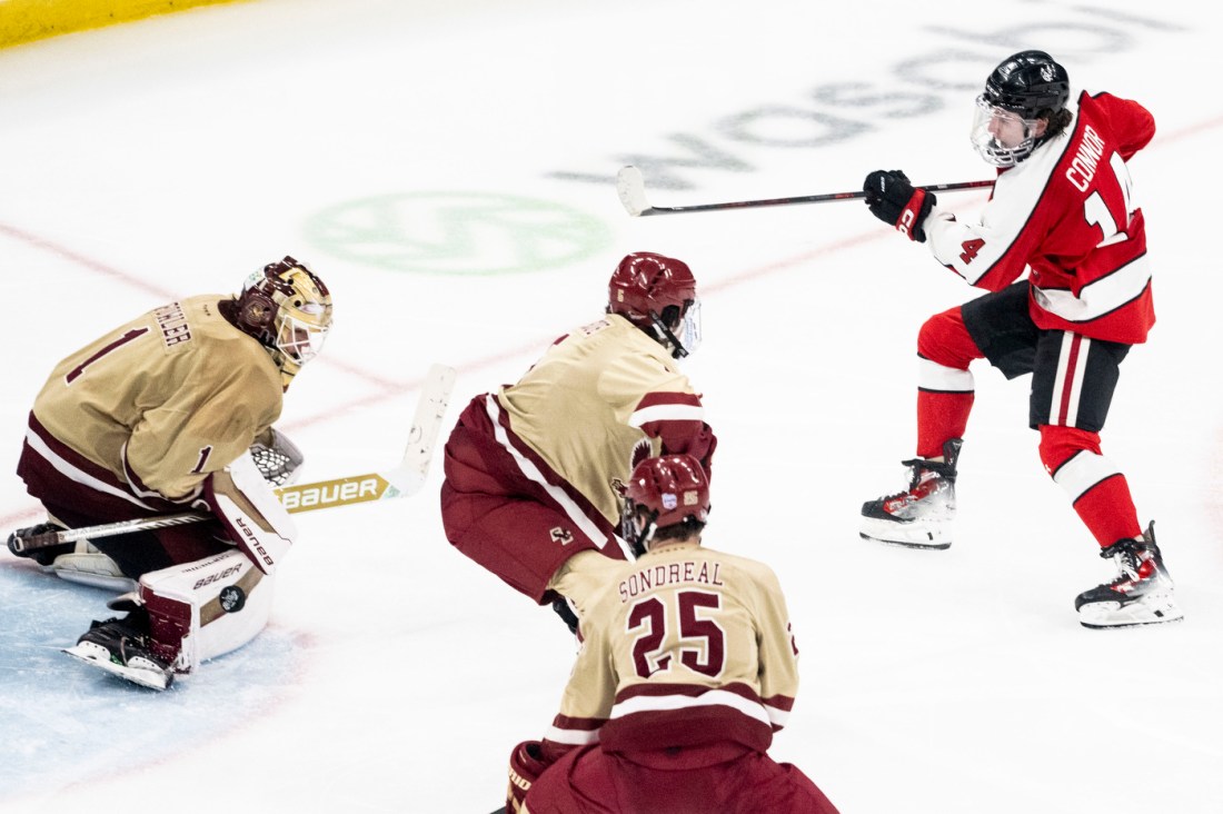 A hockey player in a red and black jersey winding up for a shot while three opposing players, including the goalie, prepare to defend.