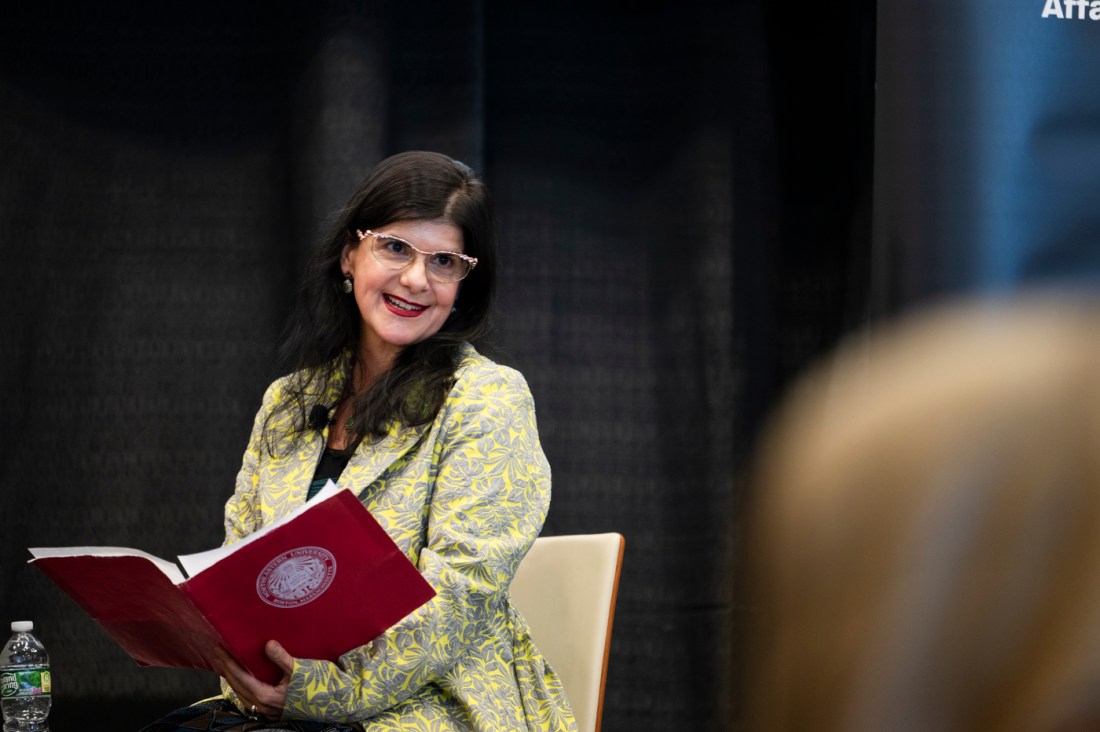 Denise Garcia wearing a green and yellow blazer and glasses sitting on stage at a fireside chat holding a book with a red cover.