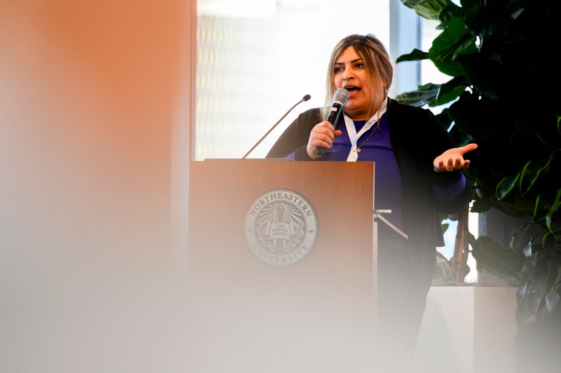 Najme Ebrahimi standing behind a podium holding a microphone in one hand and gesturing with the other. 