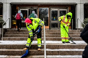 Workers in high-visibility gear shovel snow from campus steps as students walk inside.