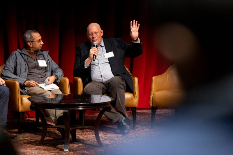 Usama Fayyad and Vinay S. Rao sitting in tan leather chairs. They are both wearing name tags. Usama Fayyad is holding a microphone in one hand while speaking into it and gesturing with his other hand.