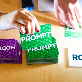 A person's hands picking up a card from a stack of cards labeled 'Prompt'. They're sitting next to a stack of cards labeled 'Modifier'.