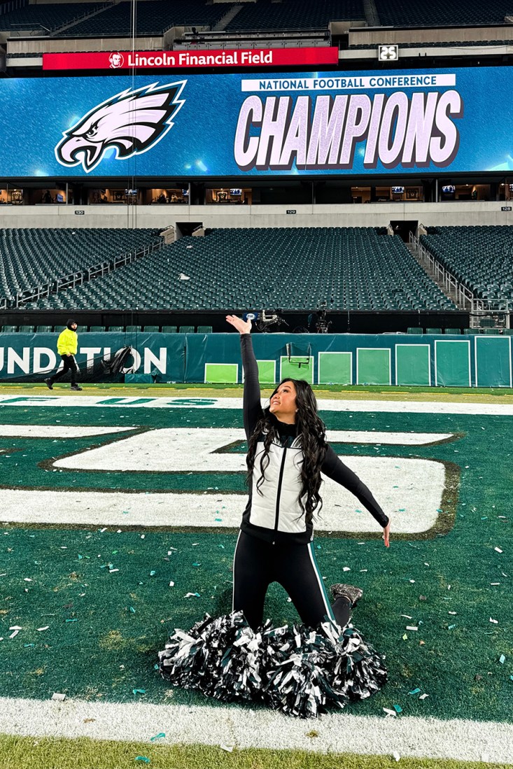 Chelsea Pe Benito kneeling on the Eagles field in Philadelphia with her pom poms in front of knees.