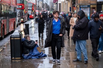 A man sitting on the pavement begging for money while shoppers walk past on a busy sidewalk.