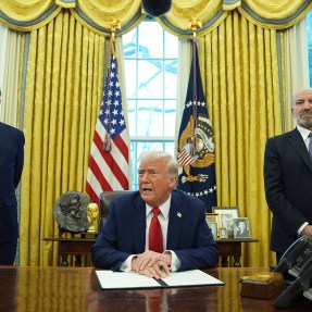 President Donald Trunp speaking while sitting in the Oval Office. Secretary Scott Bessent and Howard Lutnick stand on either side of him listening.