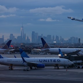 Two airplanes on a runway. In the background, a third airplane is taking off in front of a city skyline.