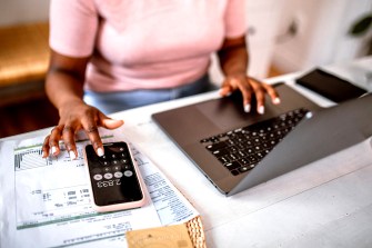 A person wearing a pink shirt sitting at a table with a laptop and their phone open to the calculator app.