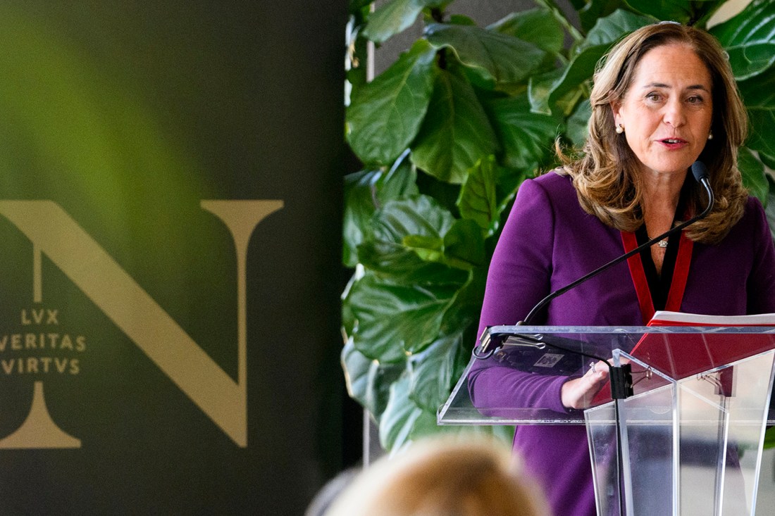 Ruth V. Aguilera wearing a purple blazer, standing at a glass podium in front of a backdrop with the Northeastern logo in gold on it.