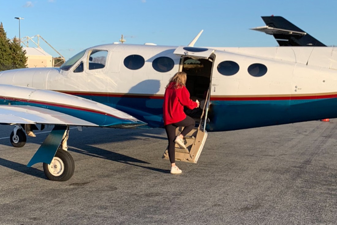 Calli Colvin wearing a red sweatshirt and black leggings stepping up into a small plane. 