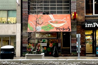 A shop on a street in Toronto with a reddish orange overhang that has a painting of a bowl of soup and the restaurant name 'Ravi Soups' on it.