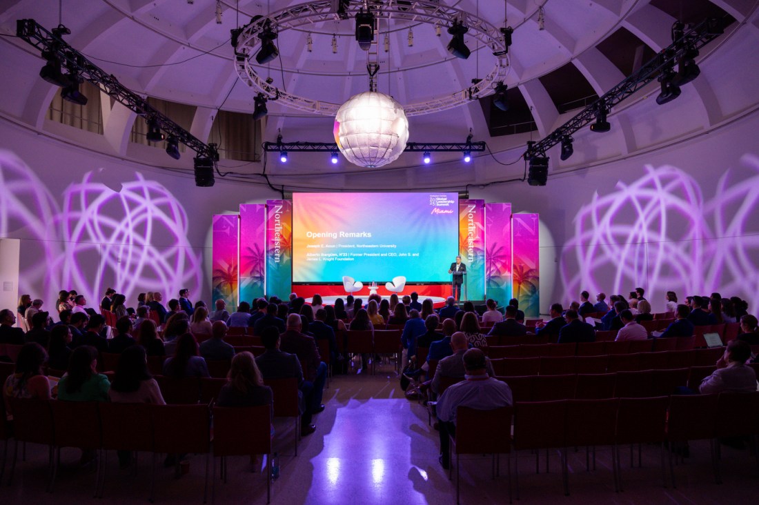 Two rows of audience members sit on either side of a walkway in a oval shaped room. At the front of the room is a stage in front of a screen. Hanging from the ceiling is a disco ball.