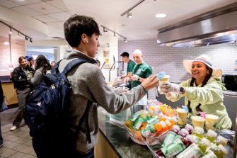 A person hands a beverage to a student at a food service counter, with other people visible in the background.