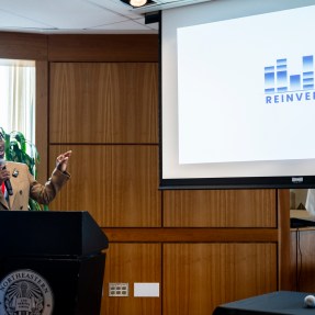 Gabby Aidam speaking in a microphone in front of a slide.