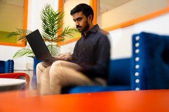 Troy Gunawardene sitting on a blue chair with his laptop in his lap as he types on it.