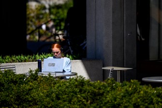 A person sits alone at an outdoor table, working on a laptop in a sunny area near a building, surrounded by greenery and shadows.
