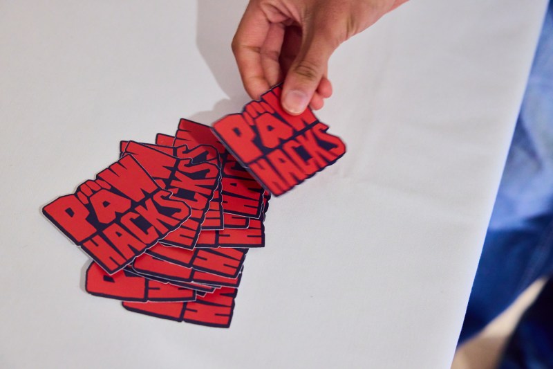 A hand picking up a sticker that says 'Paws Hacks' from a pile of stickers.