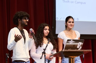 Three students standing on a stage around a podium, one of them speaking into a microphone.