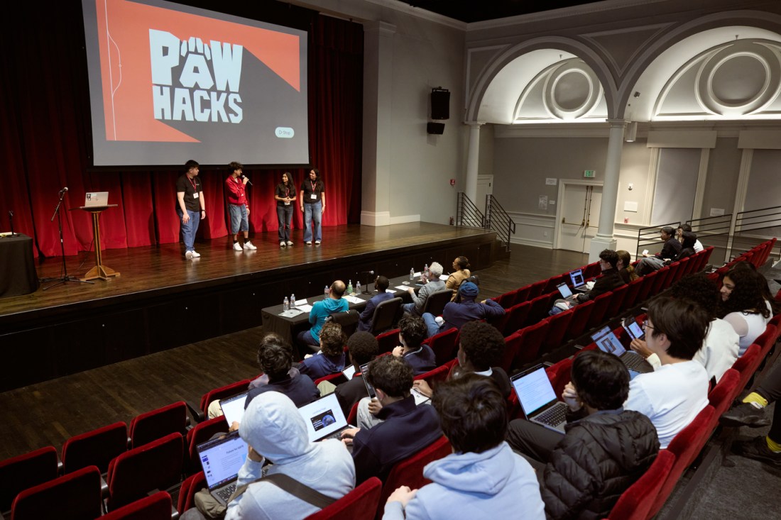 Four students standing on stage in front of a screen, one of them holding a microphone. They are at the front of a room. In front of the stage is a table with four judges. Behind the judges are rows of audience members in the auditorium chairs. 