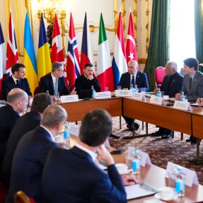 World leaders sitting around a square shaped table at a summit. Next to the table are a series of flags representing the countries that the leaders represent.