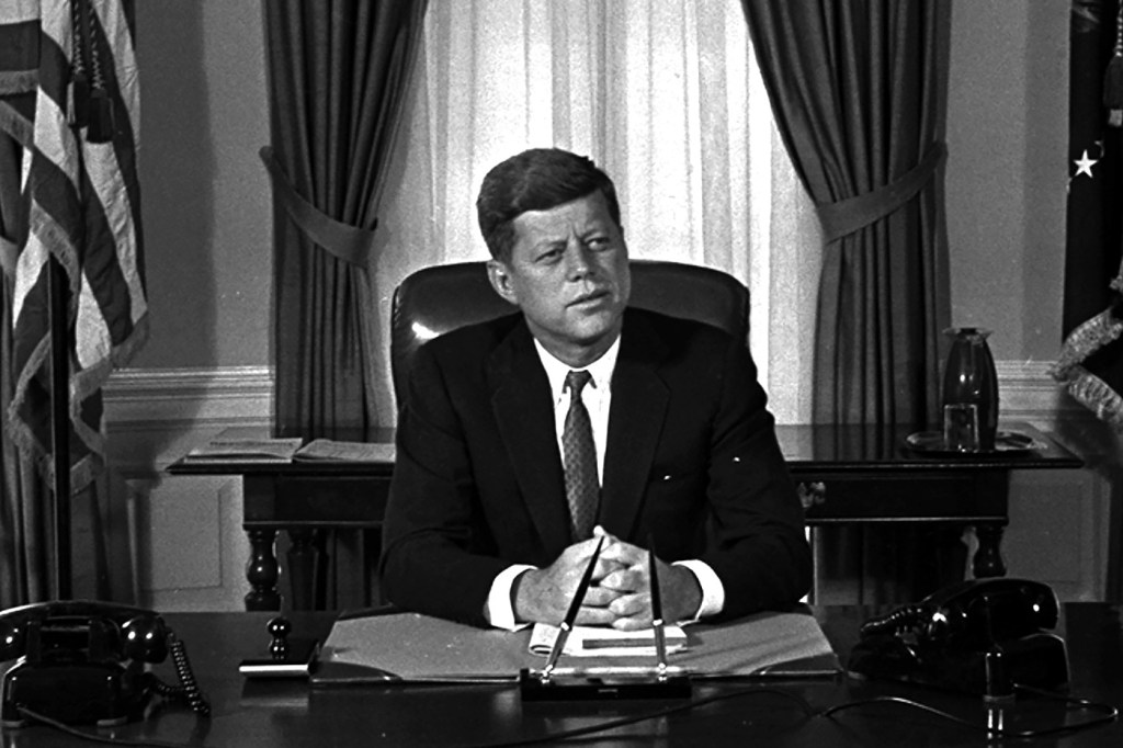A black and white photo of President Kennedy sitting at a desk at the White House.