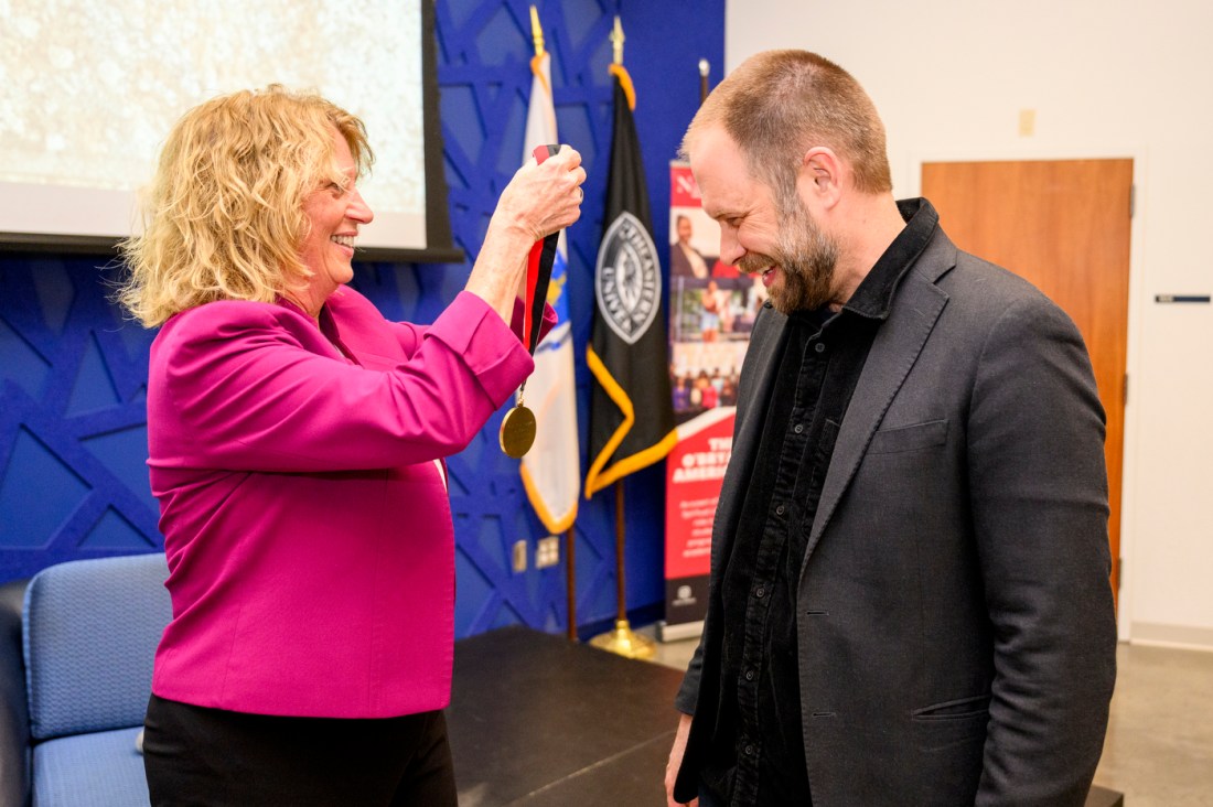 A person wearing a pink blazer placing a medal around Dan Adams' neck at the Klein Lecture.