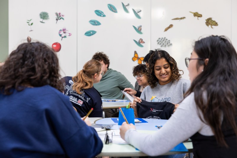 Students sitting in a room next to white boards with images of the butterfly, frog, and apple life cycle on them.