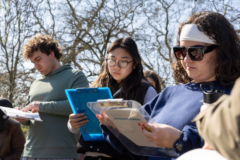 Three students sitting in Hyde Park writing on clipboards.