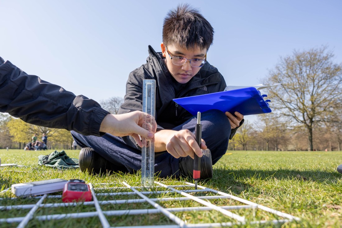 A student doing an environmental study of the grass in Hyde Park, holding an instrument in one hand and a clipboard in the other.