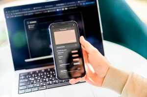 A hand holding a phone up in front of a laptop. The phone is open to an app that could help small coffee shop business owners.