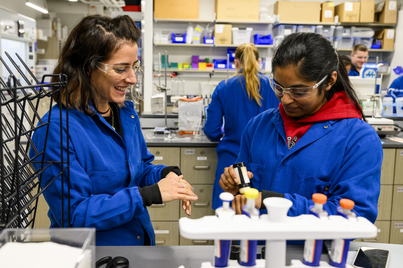 Two students wearing blue lab coats and safety glasses working on cosmetics in a lab.