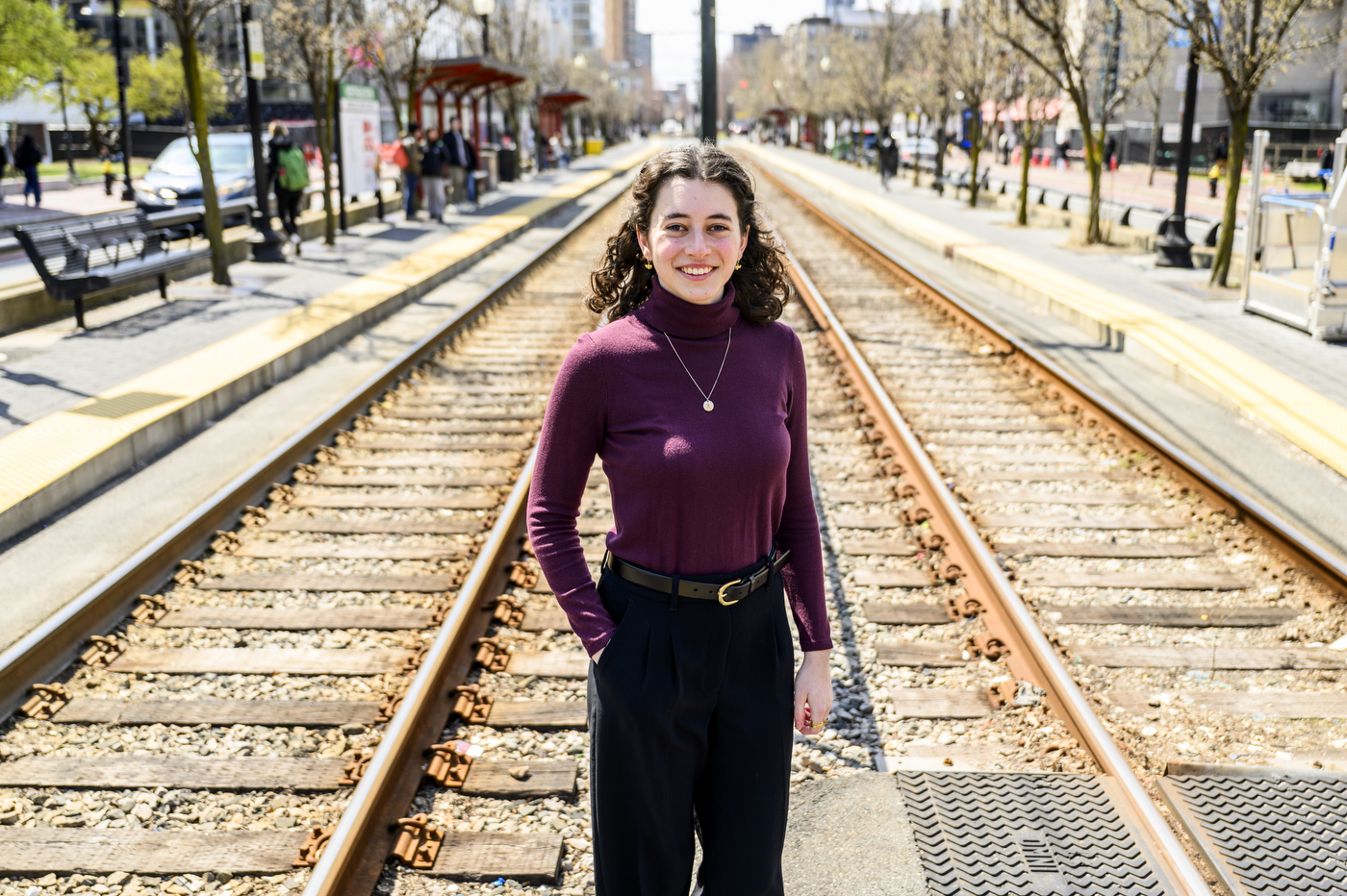 Portrait of a person standing on the green line T tracks wearing black pants, a belt, a purple turtleneck, and a necklace.