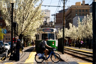 A person rides a bike across train tracks as a streetcar approaches, with blooming trees and city buildings in the background.