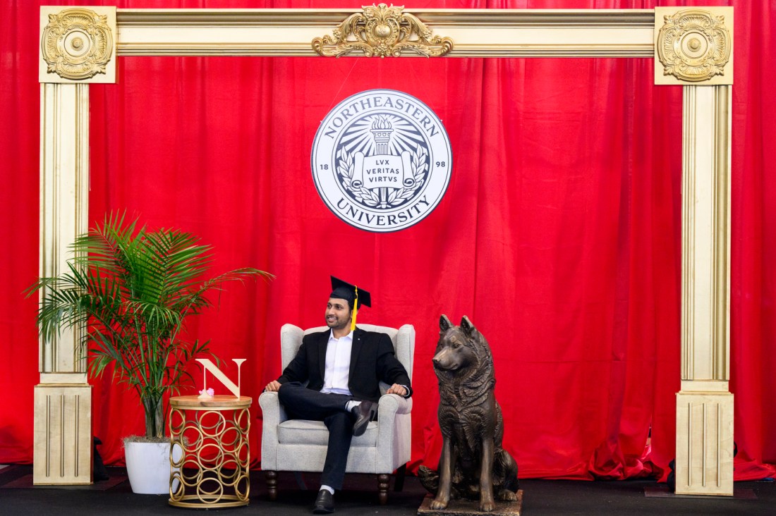 A graduate wearing a cap and gown sitting in an armchair in front of a red curtain with the Northeastern emblem above him. Next to him is a husky statue.