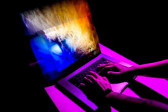A person's hands typing on a laptop in a dark room with purple lighting.