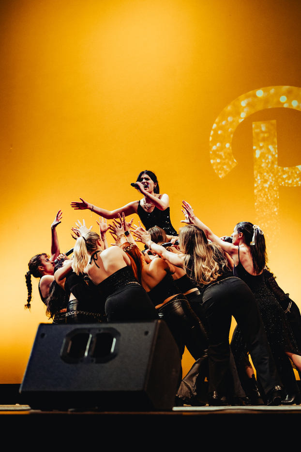 A singer from Pitch Perfect A Capella singing into a microphone surrounded by other singers from the group on stage.
