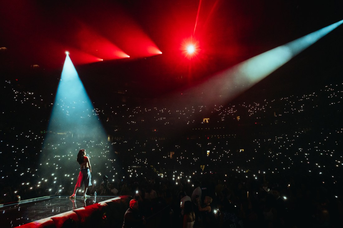A concert photo of an artist stands in spotlight onstage with crowd lights filling the arena.
