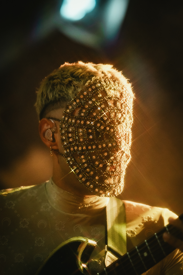 Artist performs wearing a jeweled mask and playing guitar under stage lights.