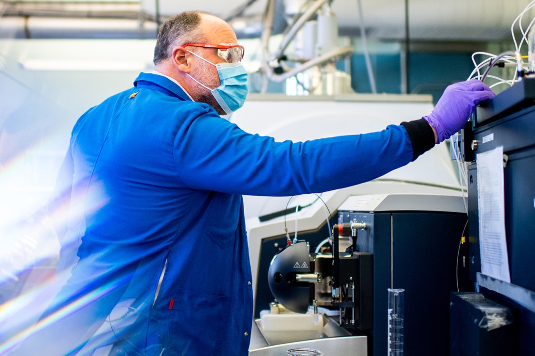 Jeffrey Agar wearing a mask, blue lab coat and purple gloves working in the lab.