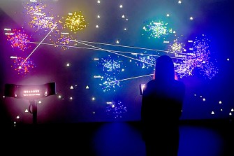 A person standing in front of an exhibit. The exhibit is a dark screen with colors mapped on to it to show people's energy impact.