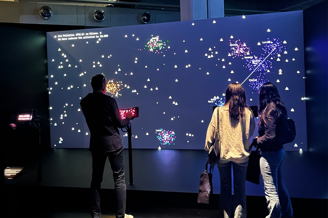 Three people standing in front of an exhibit on a screen showing a dark background with small yellow triangles and other shapes mapped on to it.