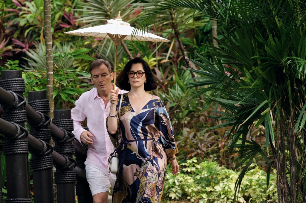 A screen capture from the "White Lotus" show that shows a character carrying a parasol walking while another character tries to keep up behind them.