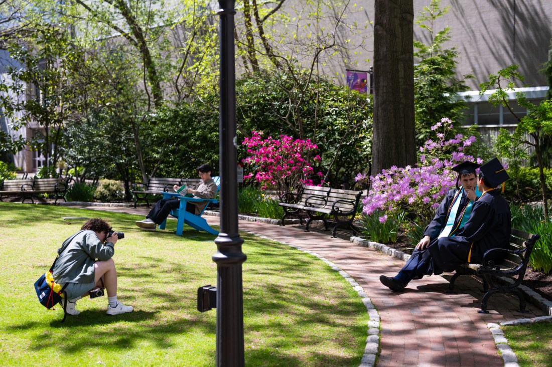 A photographer crouching to take a photo of two Northeastern graduates sitting on a bench talking to each other.