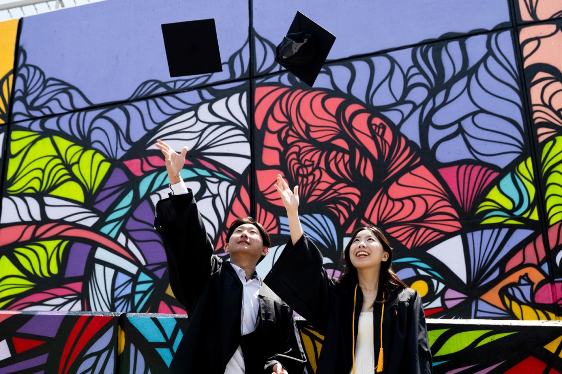 Two Northeastern graduates tossing their caps up in the air in front of a colorful mural.