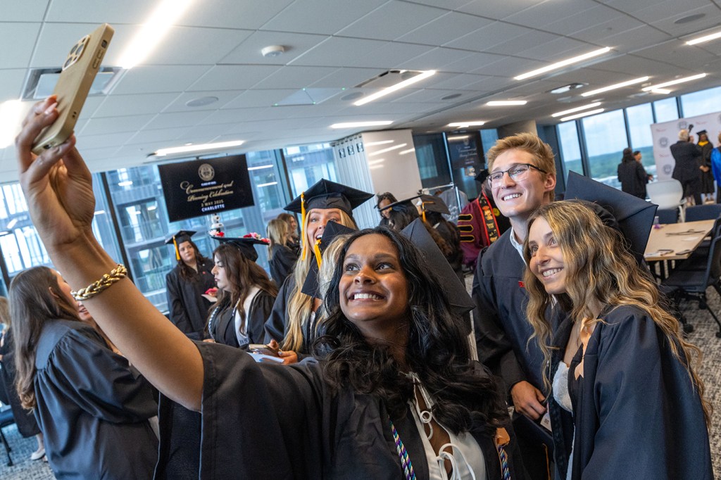 A group of people in graduation gowns smile and pose for a selfie in a bright, indoor setting.