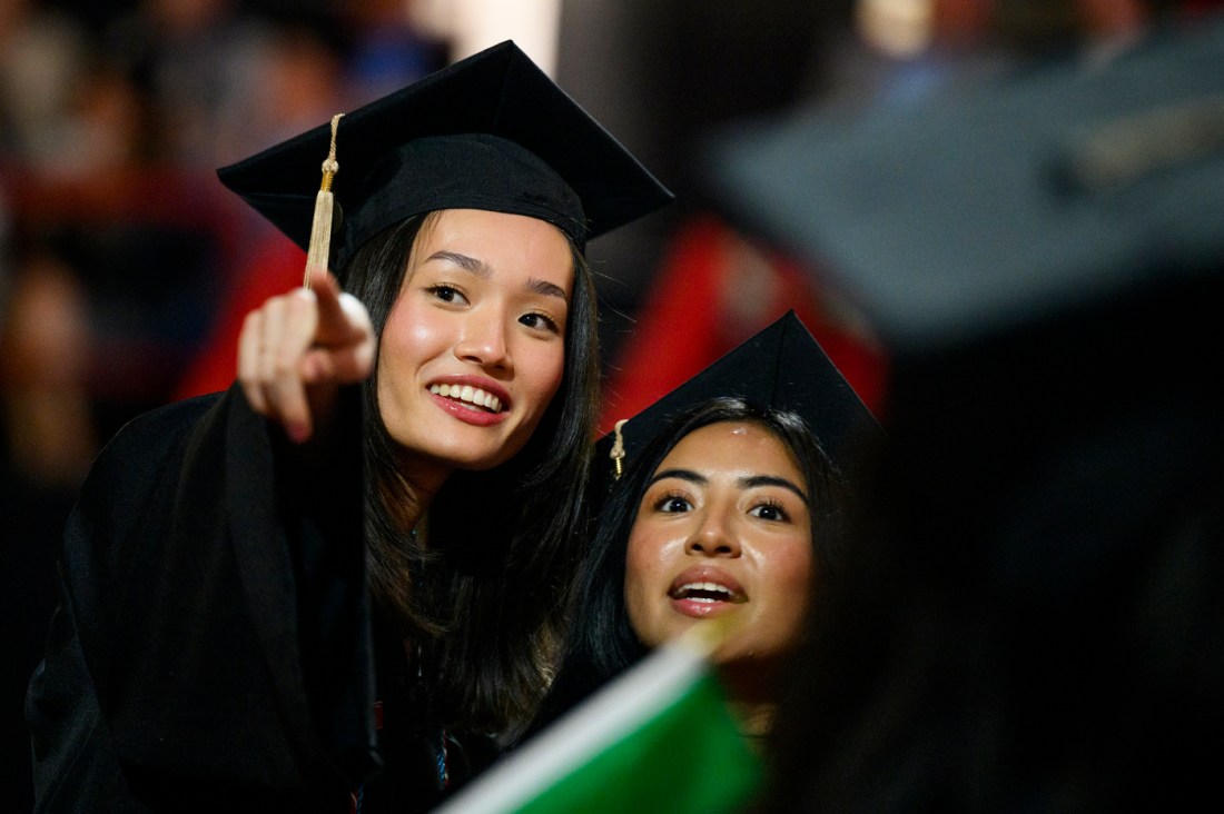 A graduate in cap and gown smiles and holds up diploma cover during a ceremony.