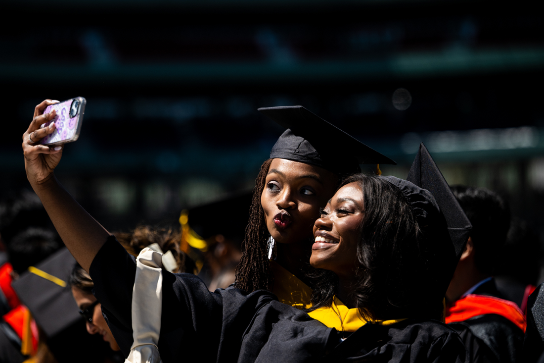 Two Northeastern graduates in caps and gowns take a celebratory selfie during Commencement at Fenway Park.