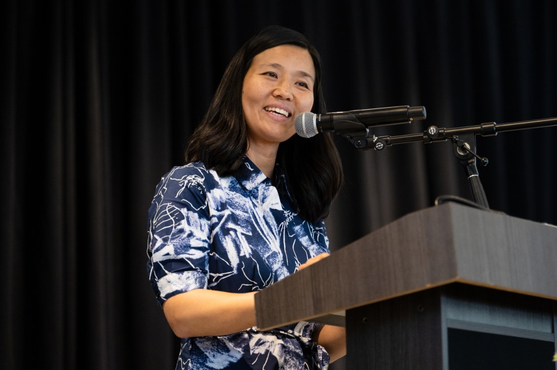 Boston Mayor Michelle Wu stands at a podium wearing a blue and white patterned dress and addressing the crowd during the Boston Public School Valedictorian Luncheon.