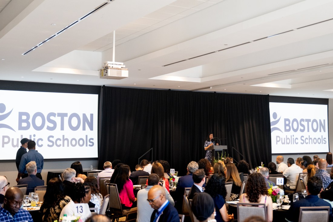 A wide shot of the audience seated at the Boston Public School Valedictorian Luncheon, with a 'Boston Public Schools' banner displaying on two screens in the background.